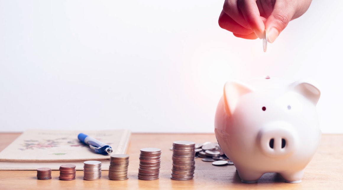 Piggy bank with coins next to it and a hand putting a coin in the piggy bank