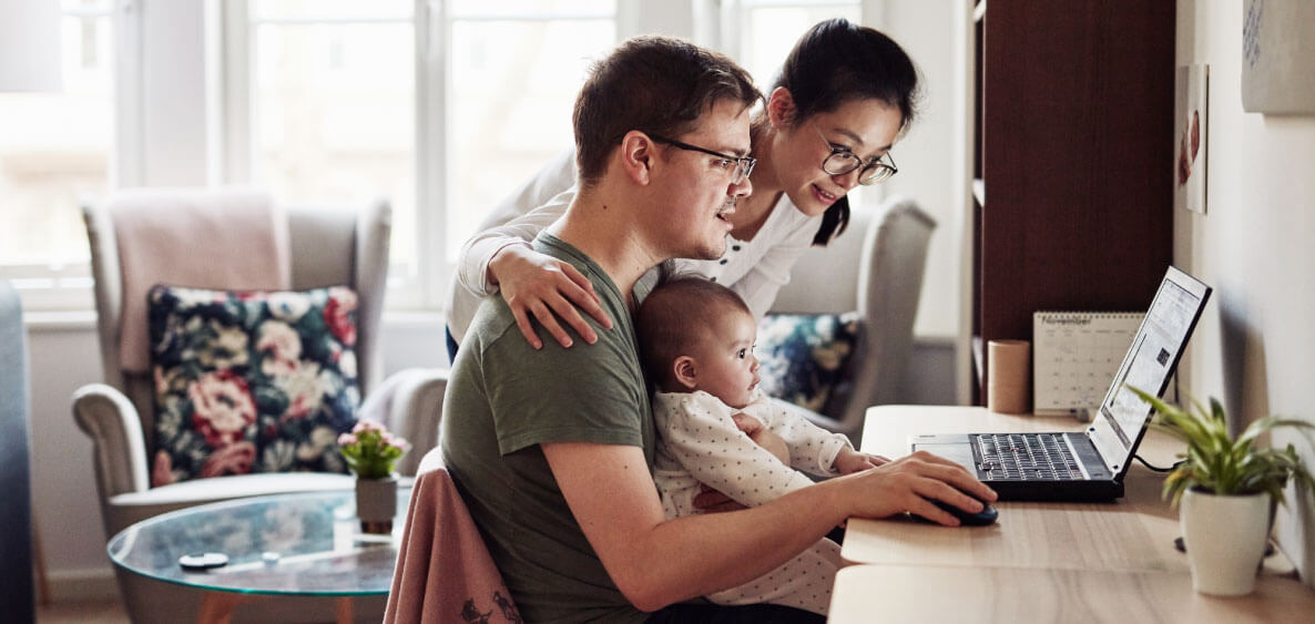 Man at his desk with a laptop with his wife on his side and baby on his lap.