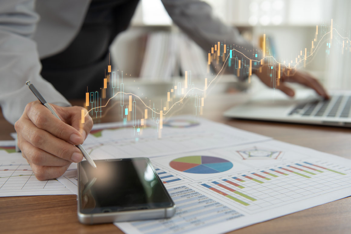 Business person has one hand holding a pen on cellphone and the other on a laptop with graphs and charts on the table.