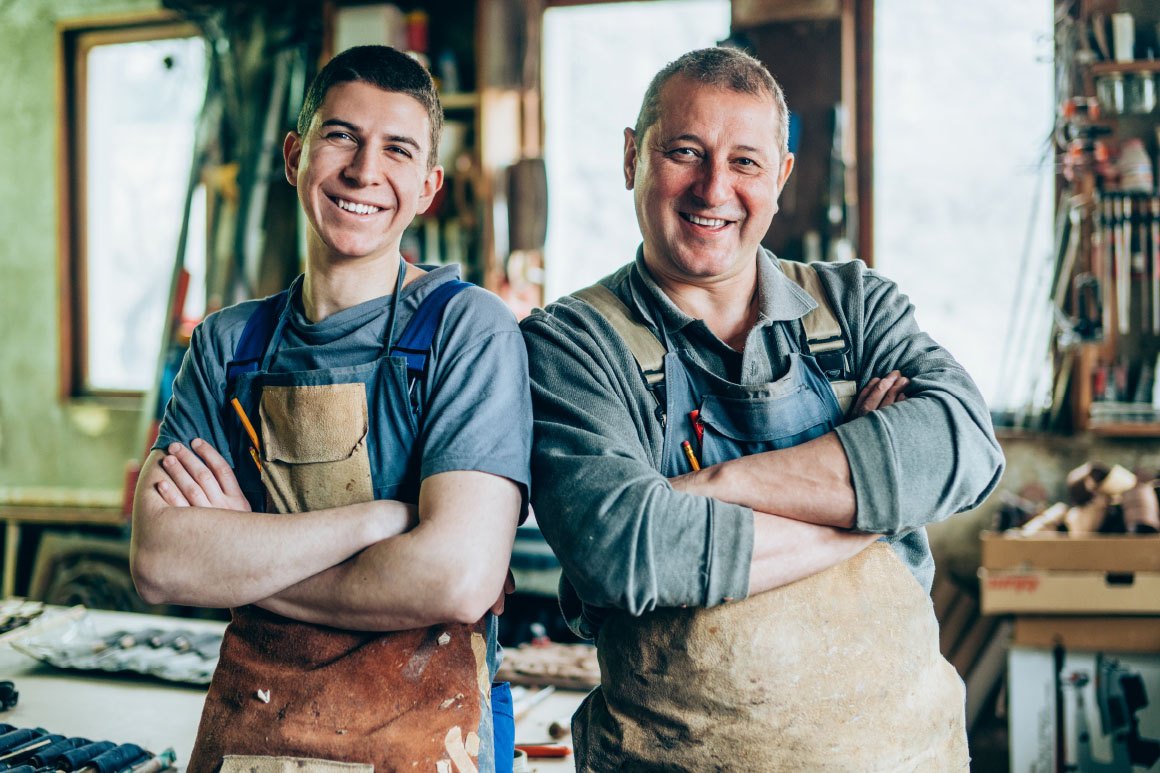 Father and son in aprons at their business smiling with their arms crossed.