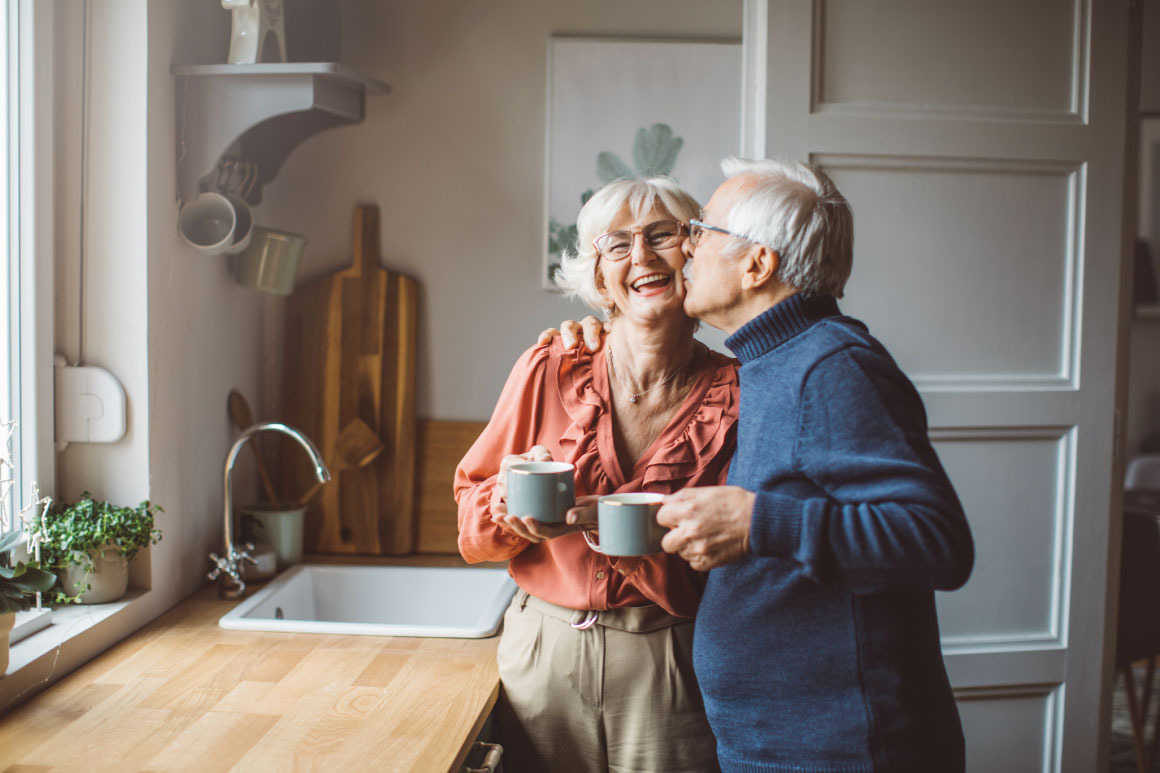 Elderly man kissing his wife on the cheek in their kitchen.