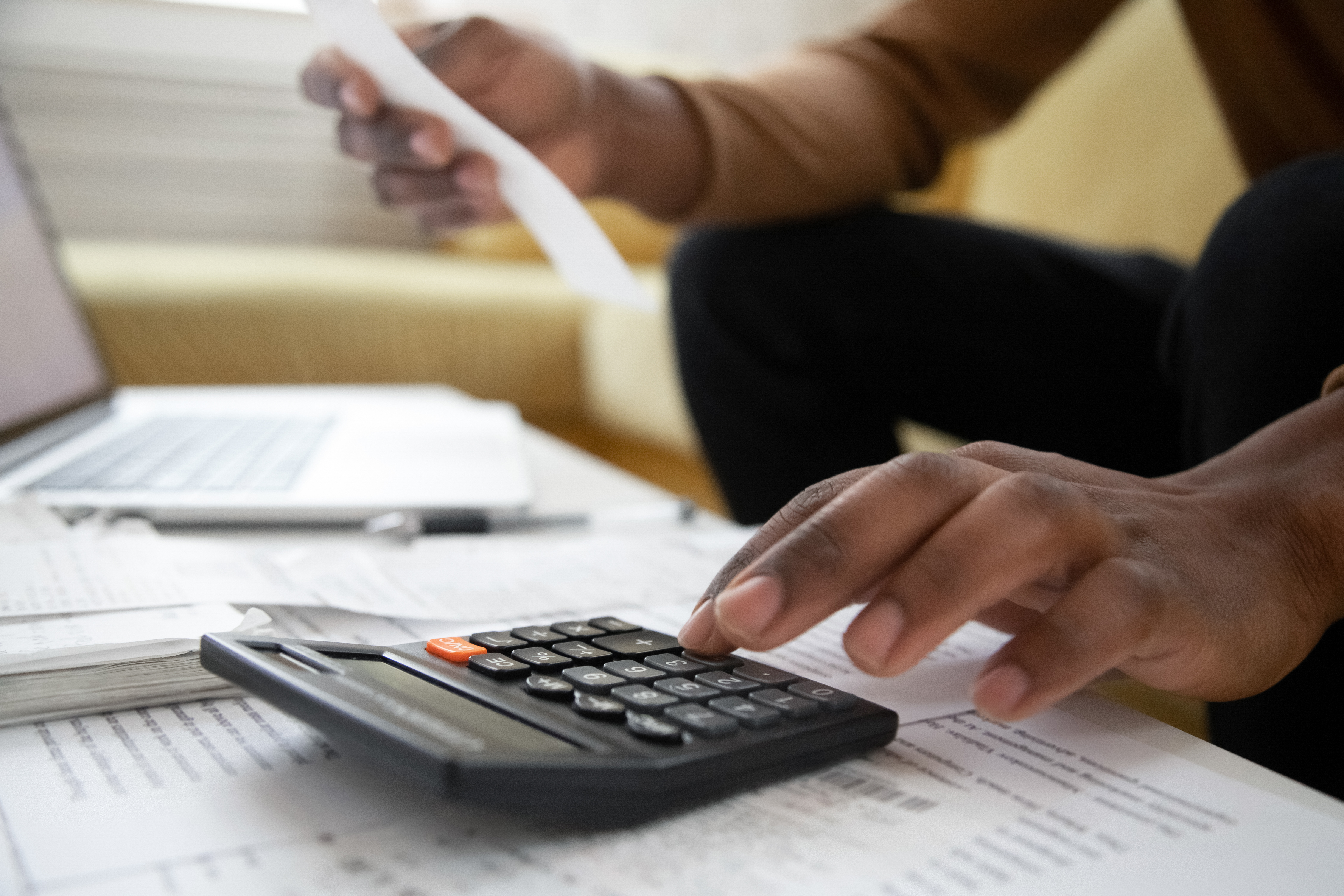Hand on a calculator with laptop open and papers on the desk