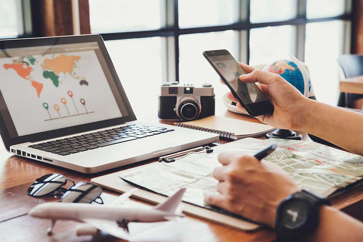 In front of bright windows, a person looks at world maps on their mobile phone and laptop while holding a pen over a paper map at a desk laden with mostly travel-related objects, including dark sunglasses, a silver and black camera, a small globe, a spiral notebook, and a 3D model of an airplane.
